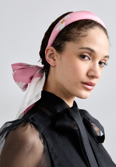 Pink silk headband adorned with white daisy patterns. Model wears a black sheer blouse with a bow tie collar. Smooth textures, elegant design.