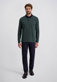 Groene polo met lange mouwen en marineblauwe kraag, voorzak en gestructureerde stof. Gecombineerd met marineblauwe broek en zwarte schoenen.