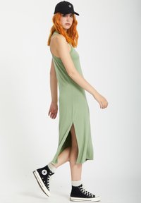 Lys grøn, ærmeløs kjole med sideslids; båret med sorte sneakers og en sort cap. Blødt stof og tætsiddende design.