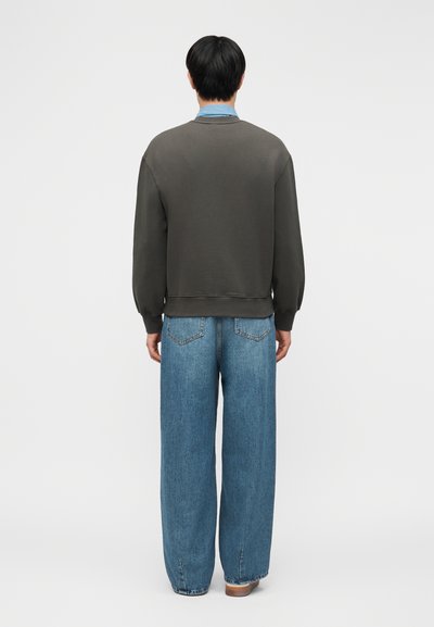 Asmuo, atsuktas nugarą, dėvi tamsiai pilką džemperį ant šviesiai mėlynos spalvos marškinio ir plačių mėlynų džinsų, rudais batais, ant balto fono.