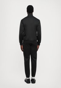 Man standing with back to camera wearing black tracksuit jacket, matching joggers, white socks, and black sneakers against white background.
