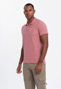 Vaaleanpunainen polo-paita, jossa on kauluspaita, kaksi nappia ja hillitty logo rinnassa. Yhdistetty beigen värisiin cargo-housuihin, joissa on taskuja.