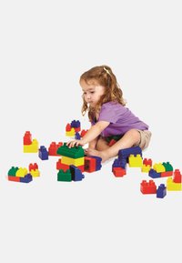 Young child sitting on the floor assembling colorful large plastic building blocks, focused on stacking a green block on a yellow-red structure.
