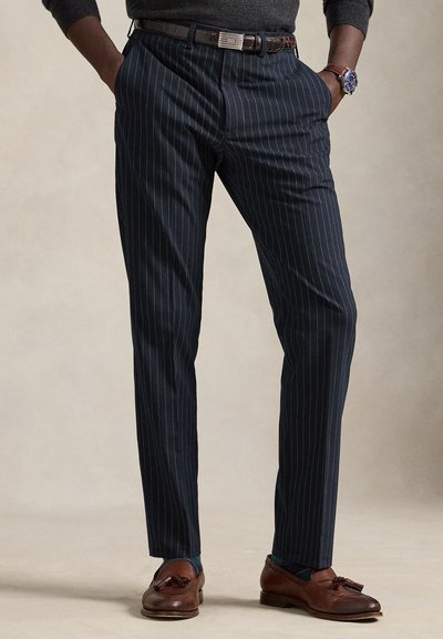 Man wearing dark pinstripe trousers, brown leather belt, brown tassel loafers, and a watch standing with hands in pockets.