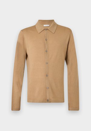 Junger Mann mit lockigem Haar steht mit den Händen in den Taschen, trägt einen beigefarbenen Knopf-Cardigan, weißes Hemd, blaue Jeans und weiße Turnschuhe.