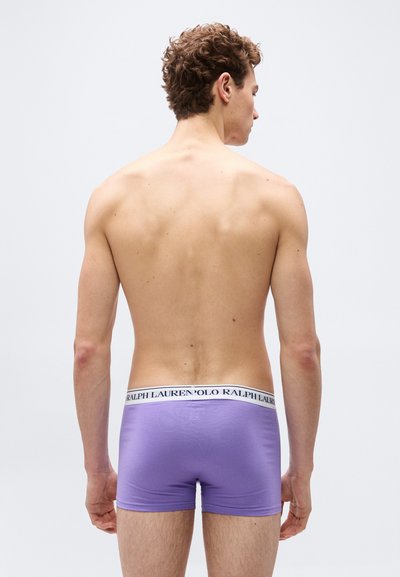 Νεαρός άνδρας με σγουρά μαλλιά, ο οποίος βλέπει από πίσω, φορώντας μοβ μποξέρ με λευκή μέση που φέρει την ετικέτα "Polo Ralph Lauren".