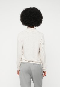 Person with curly hair wearing a light beige long-sleeve top and gray pinstriped pants, facing away against a plain background.