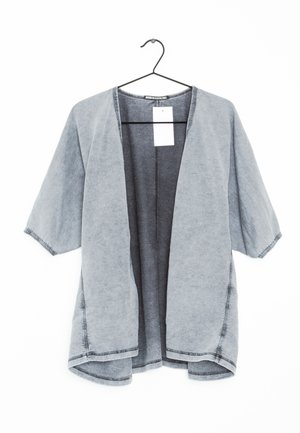 Cardigan gris clair ouvert devant avec manches courtes suspendu à un cintre noir, montrant des coutures visibles et une étiquette de prix blanche.