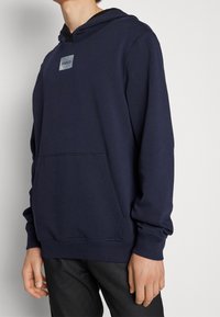 Hombre vistiendo una sudadera con capucha azul marino y un bolsillo frontal, con un pequeño logo "HUGO" en el pecho, combinado con pantalones negros.