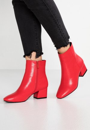 Pieds chaussés de bottines rouges vives à talons blocs et fermetures éclair latérales, associées à un jean noir coupé aux ourlets effilochés.