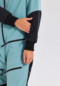 A two-tone jumpsuit in light blue and black, featuring a zip pocket on the arm, ribbed cuffs, and a textured fabric finish.