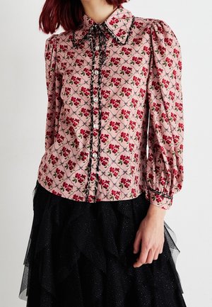 Donna che indossa una camicetta rosa con motivo floreale rosso, rifiniture in pizzo nero, maniche a sbuffo e gonna nera in tulle a balze con dettagli glitterati.