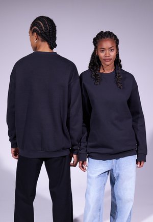 Svarte oversized hettegensere med lange ermer, rund hals og ribbestrikkede mansjetter. Modellen til venstre vender ryggen til, mens modellen til høyre vender front.