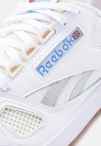 Κοντινή λήψη λευκού αθλητικού παπουτσιού Reebok, που δείχνε ι το μπλε λογότυπο με τη σημαία του Ηνωμένου Βασιλείου, λευκά κορδόνια, υφή δέρματος και αεριζόμενο δίχτυ στην πλευρά της σόλας.