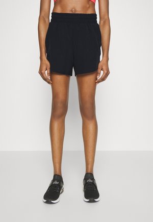 Personne athlétique portant un short noir à taille élastique, des chaussures de course noires et un soutien-gorge de sport rouge, debout sur un fond clair uni.