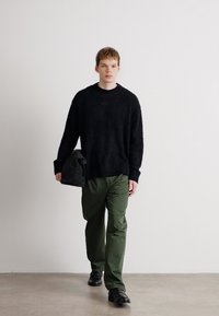 Pulover negru din tricot pufoș, pantaloni oliv și pantofi negri. Poartă o geantă neagră, mergând pe un fundal neutru.