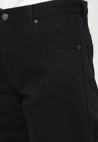 Nærbilde av sorte denimjeans med "THE JEAN MARC JACOBS" brodert på låret, som viser detaljer om lommer og knapper.