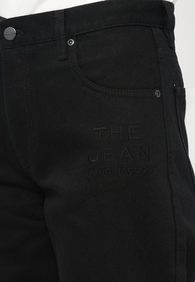 Kamerat blizu crnih traperica s natpisom "THE JEAN MARC JACOBS" izvezenim na bedru, prikazujući detalje džepova i dugmadi.