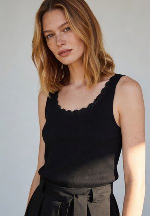 Blonde woman with freckles wearing a black sleeveless top with lace trim and high-waisted black pants with a fabric belt.