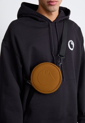 Person wearing black hoodie with white logo and round brown crossbody bag with embossed logo on front, strap across chest.