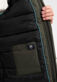 Groene en zwarte gevoerde jas binnenkant met turquoise bies, voorzien van een plat vak en een blauw label met de tekst "Industries".