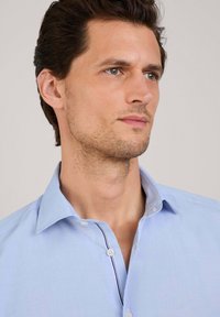 Camisa azul clara de botones hecha de una tela con textura suave. Presenta un collar clásico y un sutil acento de rayas a lo largo de la tapeta de botones.