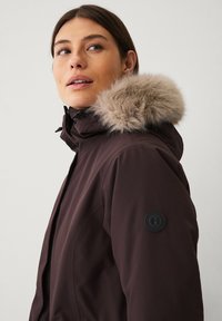 Bruine winterjas met een nepbont capuchon, ritssluiting aan de voorkant en een logo-patch op de mouw. Glad buitenmateriaal, slim fit ontwerp.
