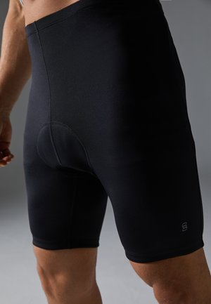 Close-up van een persoon die zwarte gewatteerde fietsbroek draagt, met zicht op de rug en bovenbenen tegen een grijze achtergrond.