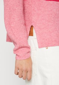 Rosa gerippter Pullover mit langen Ärmeln und seitlichen Schlitzen. Verfügt über ein strukturiertes Material und ein sichtbares Etikett am Saum, kombiniert mit weißen Hosen.