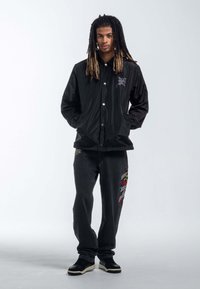Chaqueta negra y brillante con botones a presión y detalles de logo, combinada con pantalones gris oscuro que presentan diseños bordados coloridos. Zapatillas negras completan el look.