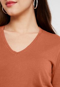 Rostfärgad t-shirt med v-hals och korta ärmar, slät textur och subtila sömnadsdetaljer. Visas på en modells halslinje.