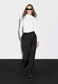 Witte langemouwen top met een capuchon, gecombineerd met hooggetailleerde zwarte geruite broeken en zwarte sandaaltjes met bandjes. Model draagt zwarte zonnebril.