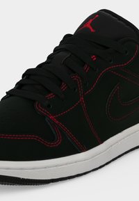 Μαύρο και κόκκινο παπούτσι Nike Air Jordan με μαύρα κορδόνια, κόκκινες ραφές και λευκή σόλα, δείχνοντας τη γλώσσα με το κόκκινο λογότυπο Jumpman.