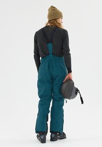 Teal snöbyxor med justerbara svarta hängslen, med en smal design på benen. En person håller en grå hjälm och bär en beige mössa.