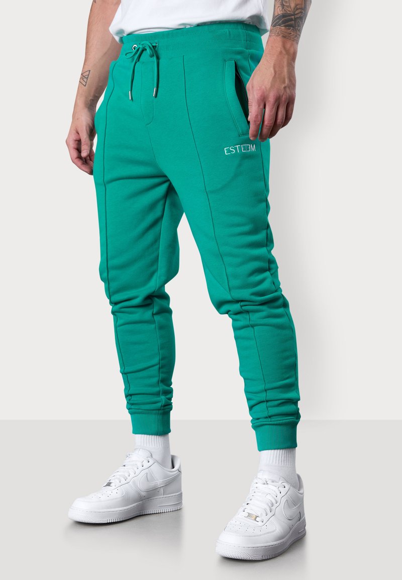Türkise Jogginghosen mit elastischem Bund, Kordelzug, seitlichen Taschen und weißem gesticktem "ESTEEM"-Logo, kombiniert mit weißen Sneakern.