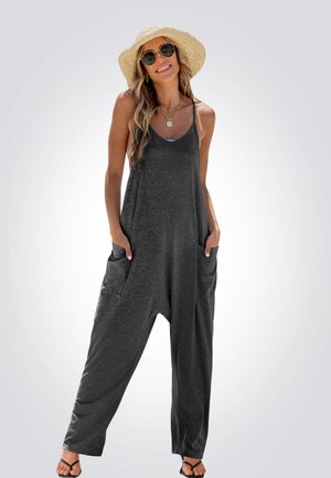 Grijze mouwloze jumpsuit met een losse pasvorm, grote zijzakken en een zachte textuur, gecombineerd met zwarte sandalen en een strohoed.