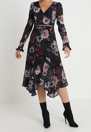 Vrouw draagt een zwarte midi-jurk met bloemenprint, lange mouwen, V-hals, asymmetrische zoom en doorschijnende stof, gecombineerd met zwarte enkellaarsjes met hoge hak.