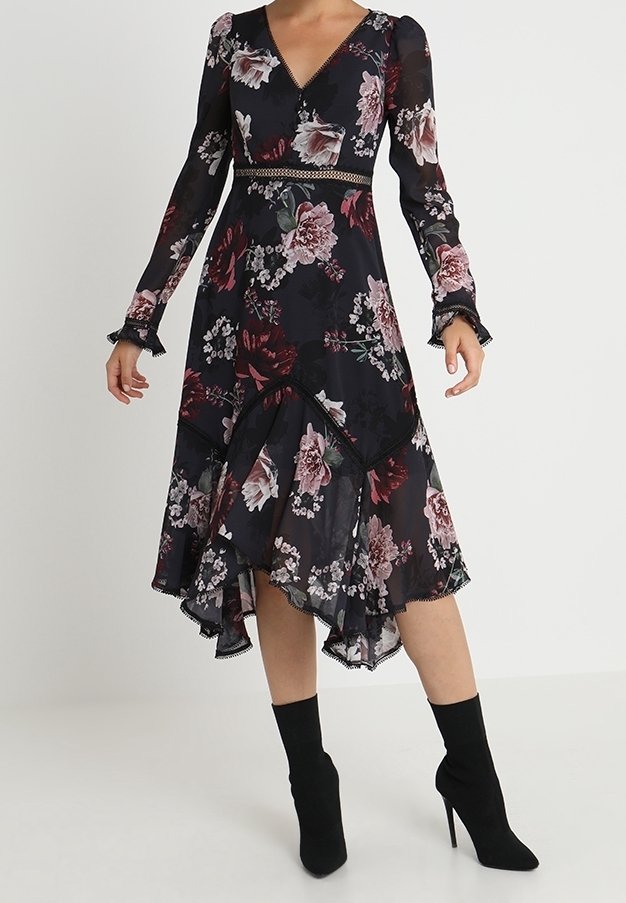 Femme portant une robe midi noire à fleurs avec manches longues, col en V, ourlet asymétrique et tissu transparent, associée à des bottes noires à talons hauts.