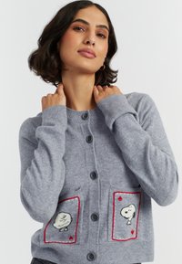 Een vrouw met donker golvend haar poseert zelfverzekerd in een grijze cardigan met speelse Snoopy-ontwerpen in de zakken, tegen een effen achtergrond.