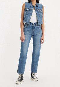 Chaleco vaquero azul con acabado deshilachado, camiseta sin mangas blanca, vaqueros azul de talle alto con tachuelas laterales y zapatillas negras con suelas y cordones blancos.