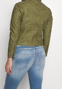 Kurtka ze sztucznej skóry w oliwkowej zieleni o dopasowanym fasonie, z pikowanymi detalami na ramionach i suwakami przy mankietach, zestawiona z jasnoniebieskimi dżinsami.
