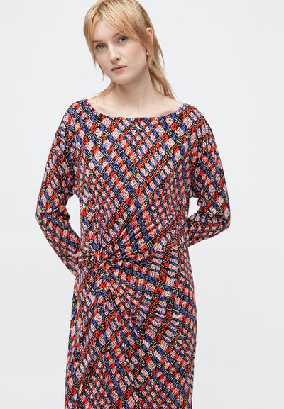 Vestido estampado multicolor com mangas longas, decote arredondado e detalhe de cintura torcida. O tecido apresenta desenhos geométricos nas cores vermelha, azul e preta.