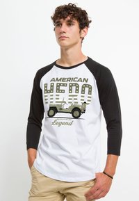 Raglan baseball t-shirt med sorte ærmer og hvidt krop. Har teksten "AMERICAN HERO", en jeep-graphic i olivengrøn og stjerner detaljer.