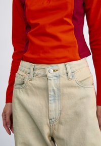 Ljusblå, högmidjade jeans med dragkedja och knappstängning; har fem fickor och subtilt blekt mönster. Kombination med en topp i röd och lila.