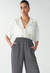 Blusa bianca in raso a maniche corte con due tasche frontali abbinata a pantaloni larghi a vita alta in bianco e nero con motivo.