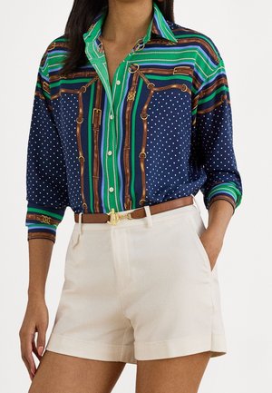 Kobieta nosząca niebieską, zieloną i brązową koszulę na guziki w białe groszki, wpuszczoną w beżowe szorty z wysokim stanem i brązowym paskiem.