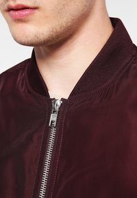 Burgunderfarbene Bomberjacke mit glatter, glänzender Textur. Verfügt über einen Reißverschluss und einen gerippten Kragen. Sichtbarer Metallreißverschluss und Nahtdetails.