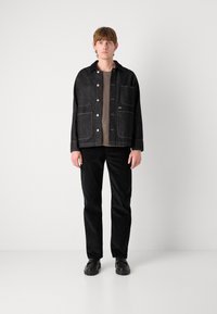 Black denim jacket with two front pockets, button closure, and a textured brown sweater underneath. Paired with black corduroy pants and shoes.