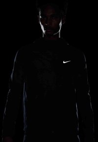 Czarna sportowa kurtka z kapturem i odblaskowym logo Nike na klatce piersiowej. Materiał wydaje się gładki, z dopasowanym krojem. Ambientowe oświetlenie tworzy sylwetkę.
