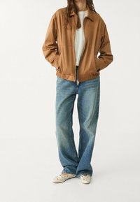 Chaqueta marrón con cremallera hecha de material suave, con cuello y cinturilla elástica, combinada con vaqueros de pierna ancha de color azul claro y zapatos de slip-on crema.